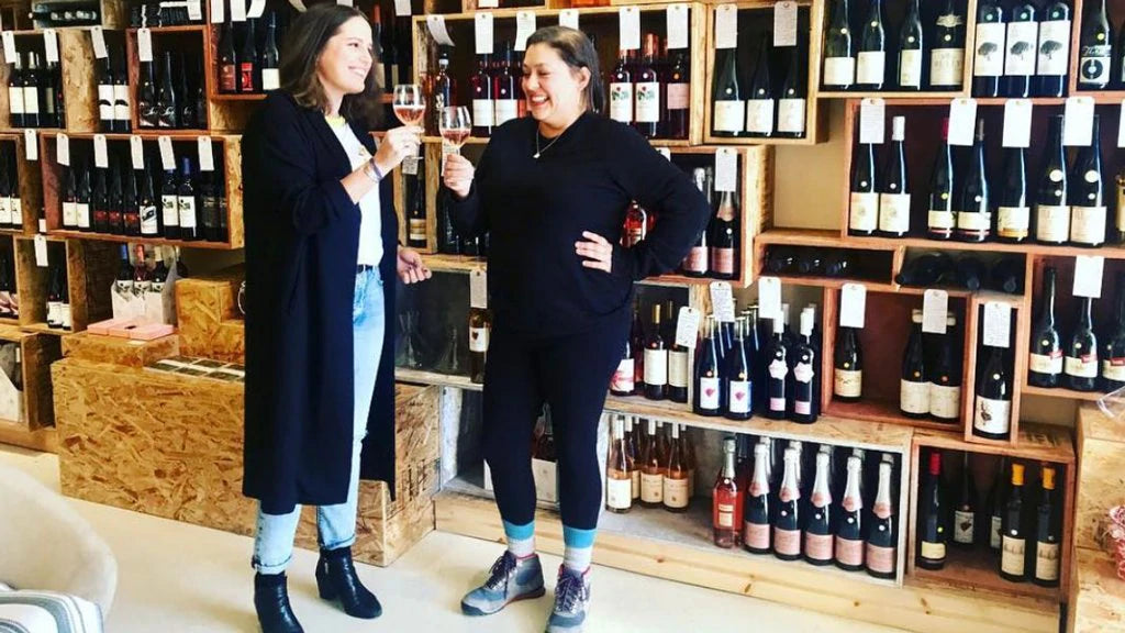 Vinovore owner Coly cheersing a glass of wine with a female patron, in front of a wall of wine on shelves