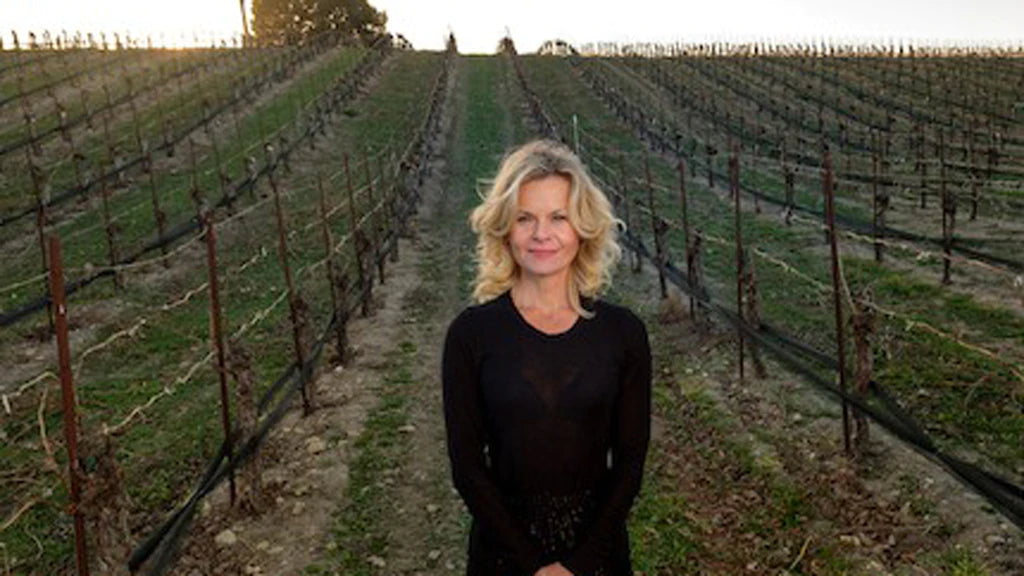 A blond woman in black standing in a vineyard with very young vines, almost no leaves