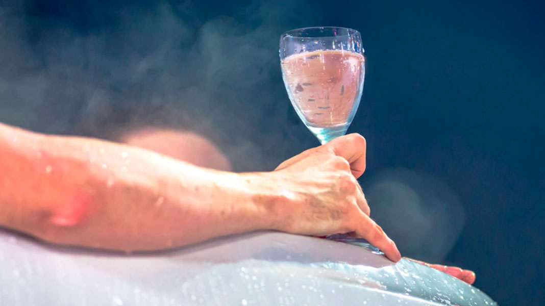 A hand holding a glass of rosé wine in a steamy tub