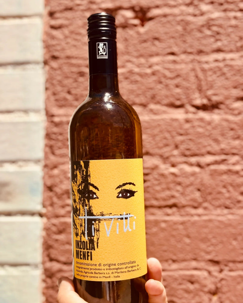 Bottle of wine with a yellow label featuring a face design, held against a brick wall.
