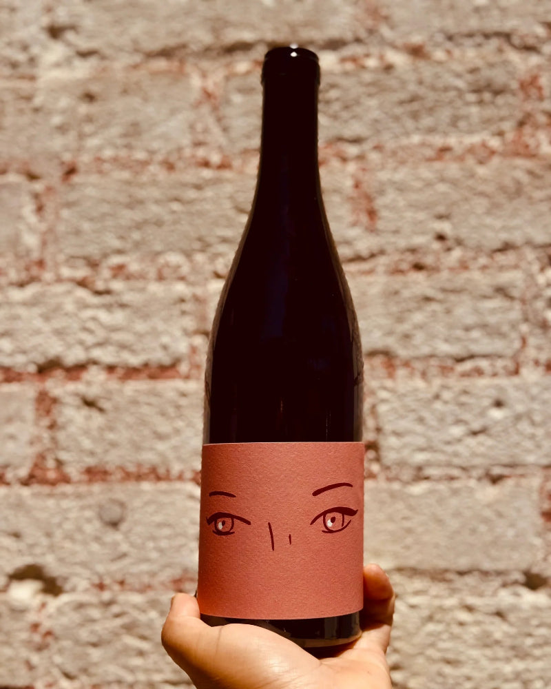 Wine bottle with decorative label featuring a face design held against a brick wall.