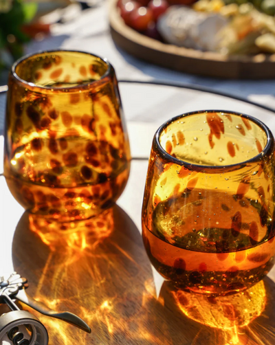 Beautiful red wine glasses add a unique touch to your table. This amber speckled stemless wine glass set celebrates the art and traditions of Mexican artisans and is hand blown from recycled glass. Quantity: 2 stemless wine glasses Materials: Recycled glass Capacity: 13 oz. Care: Hand wash recommended.   Woman Owned